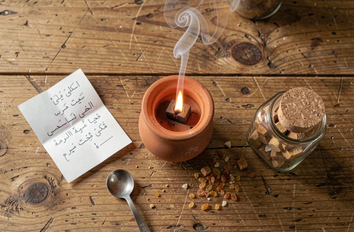 Vue aérienne d'un brûleur à encens mabkhara avec un seul morceau de bakhour dosé, entouré d'accessoires de mesure sur une table en bois brut